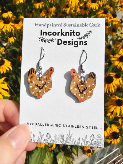 Handpainted natural cork chicken earrings on silver hooks, displayed on Incorknito Designs branded card held in hand against rudbekia