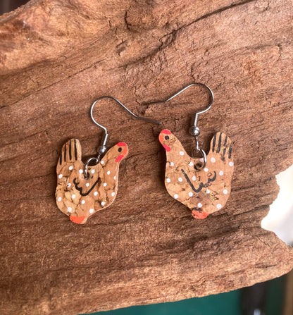 Close-up of handpainted natural cork chicken earrings on silver hooks, resting on driftwood showing cork texture detail