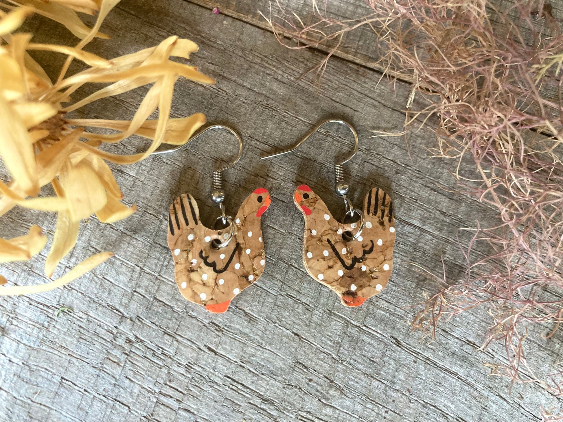 Handpainted natural cork chicken earrings on silver hooks, flat lay on weathered wood with dried botanicals