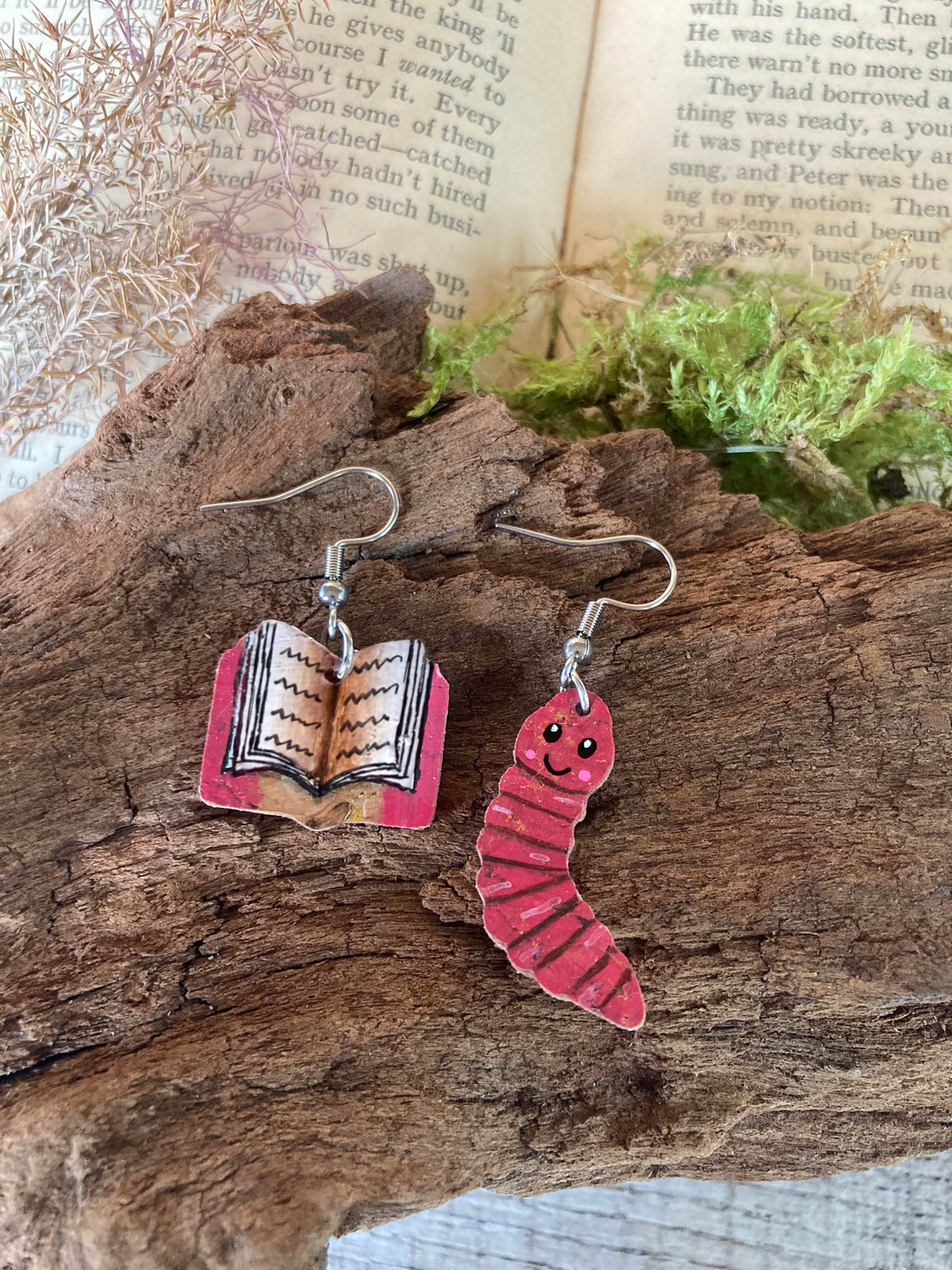 Handpainted mismatched cork bookworm earrings on silver hooks — pink open book and pink worm charms resting on cork bark with open vintage book background and moss