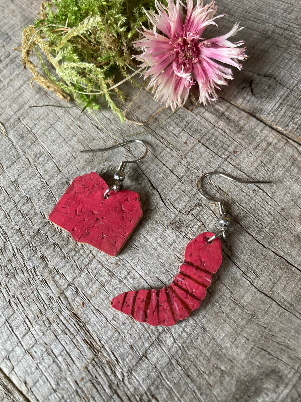 Cork bookworm mismatched earrings showing natural cork reverse — pink book and worm charms on silver hooks, flat lay on weathered wood with pink dried flower