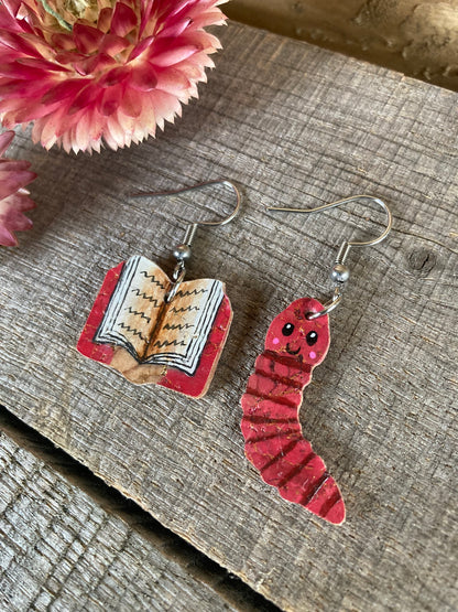 Mismatched cork bookworm earrings on silver hooks — pink open book and pink worm charms, flat lay on weathered wood with pink dried flower
