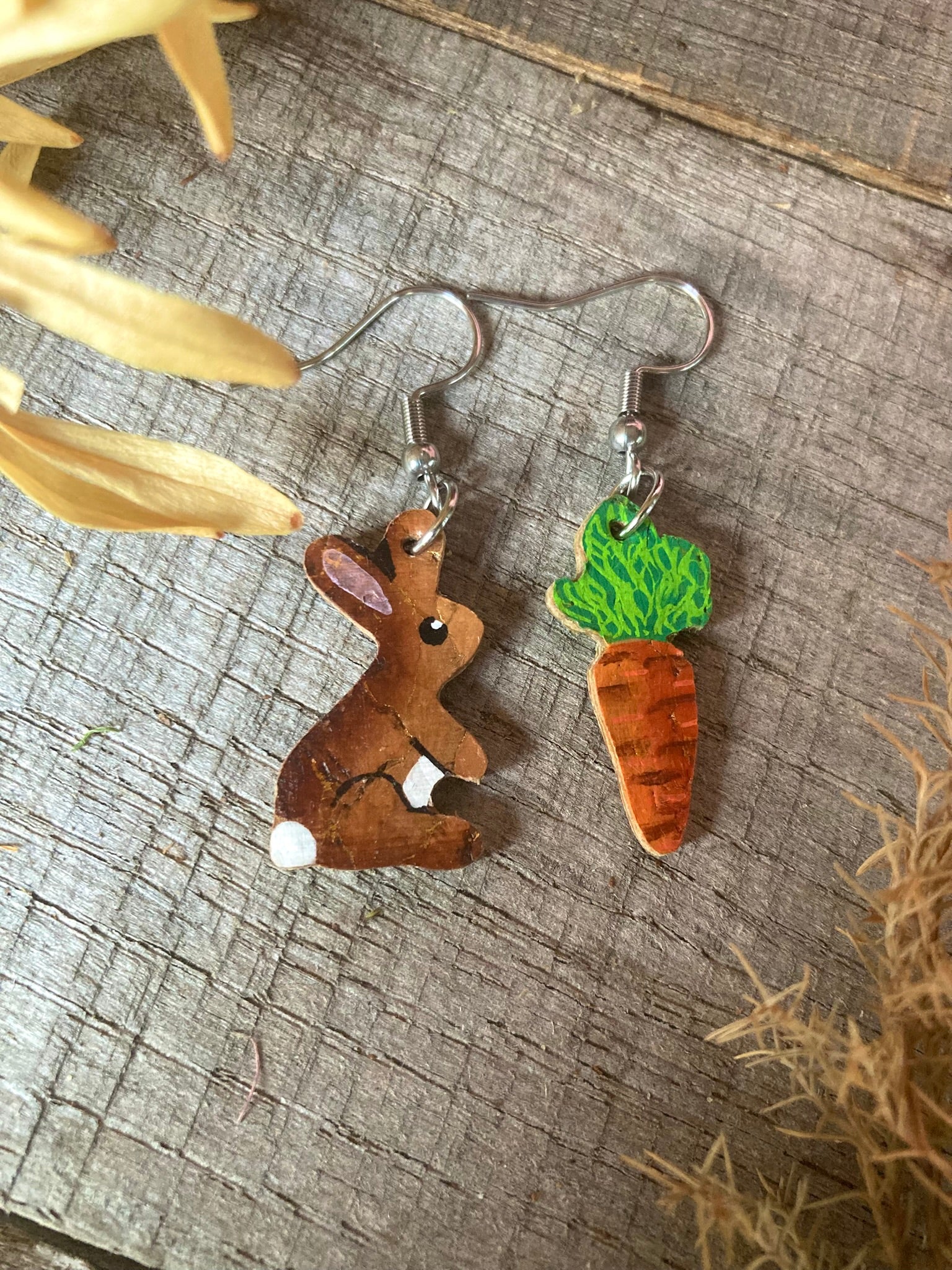 Handpainted cork rabbit and carrot earrings laid flat on a weathered wooden surface.
