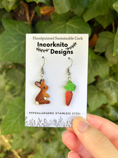 Cork rabbit and carrot hook earrings on an Incorknito Designs card, held in hand against a lush ivy background.