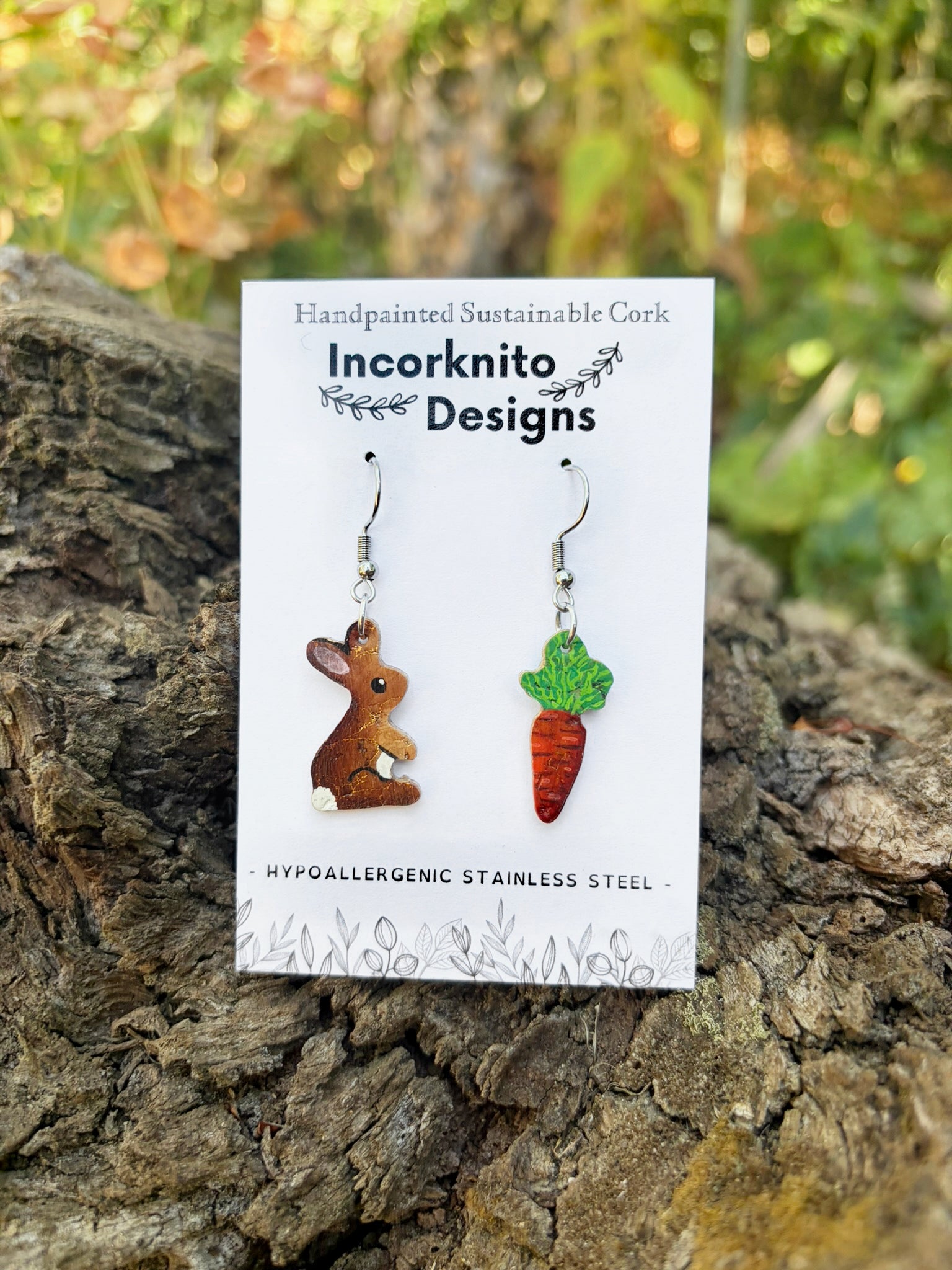 Cork rabbit and carrot hook earrings on an Incorknito Designs card, propped against a mossy log with a garden background.
