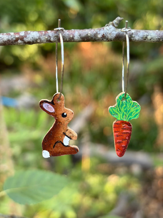 Handpainted cork rabbit and carrot hoop earrings hanging from a lichen-covered branch against a soft green garden background.