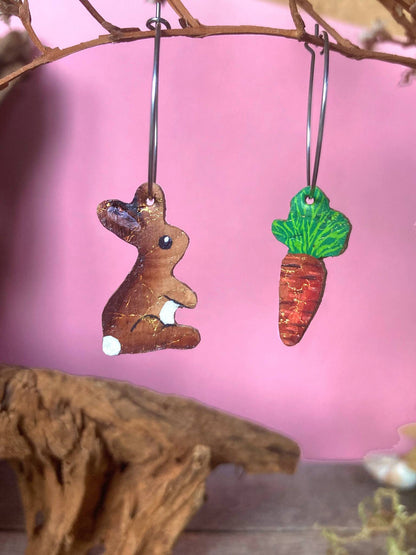 Cork rabbit and carrot hoop earrings hanging against a soft pink background.