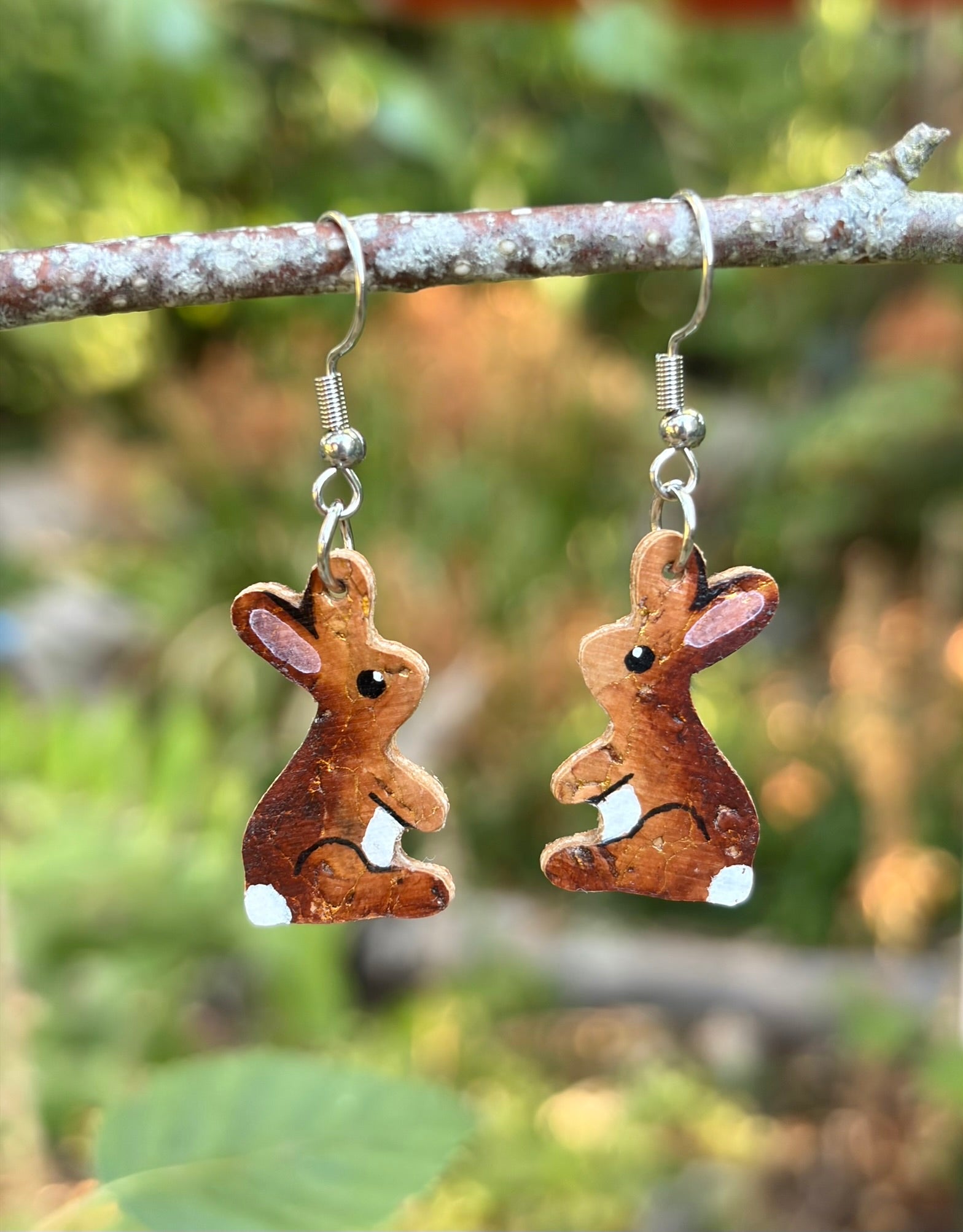 Handpainted cork rabbit hook earrings hanging from a lichen-covered branch against a soft green garden background.
