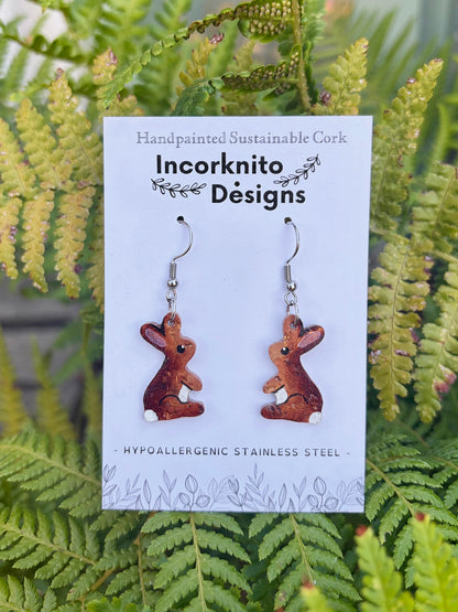 Cork rabbit hook earrings on an Incorknito Designs card, nestled amongst lush green fern fronds.