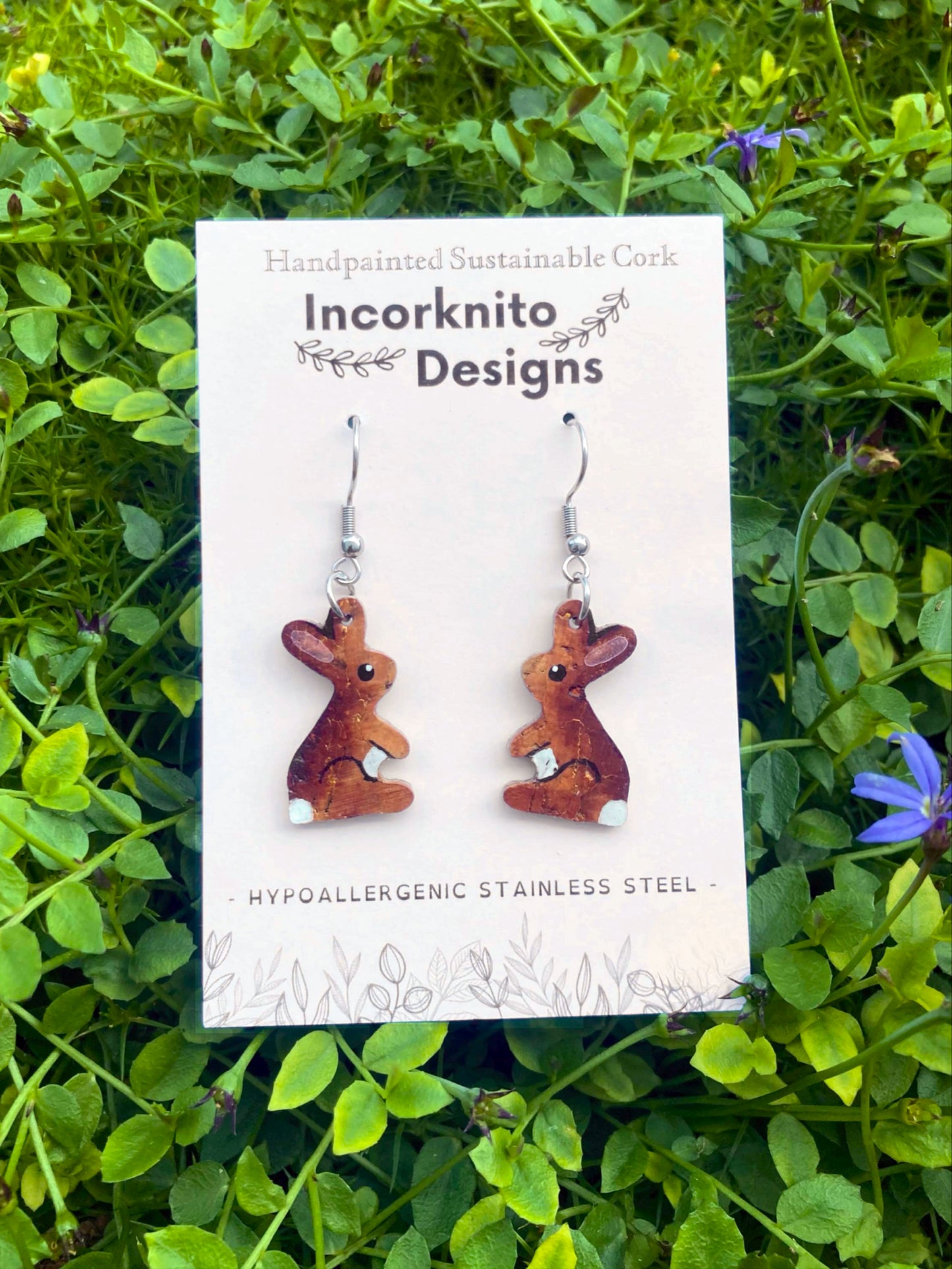 Handpainted cork rabbit earrings on an Incorknito Designs card, laid flat on a bed of wildflowers 