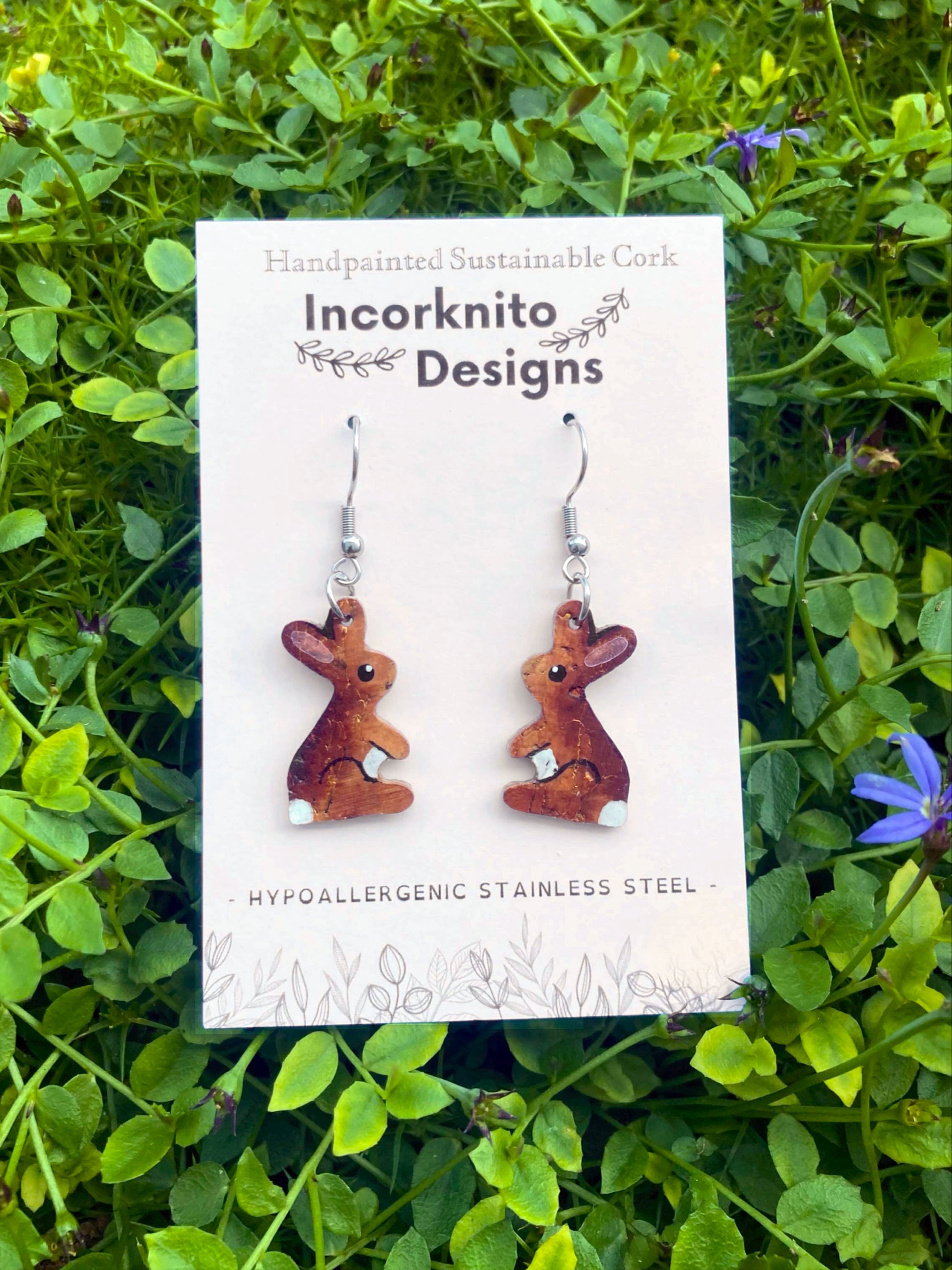 Handpainted cork rabbit earrings on an Incorknito Designs card, laid flat on a bed of wildflowers 