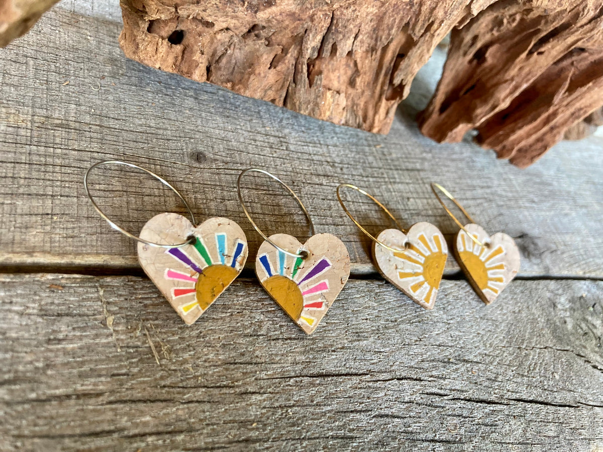 Rainbow multicoloured and yellow sun heart cork earrings on gold hoops, flat lay on weathered wood with cork bark