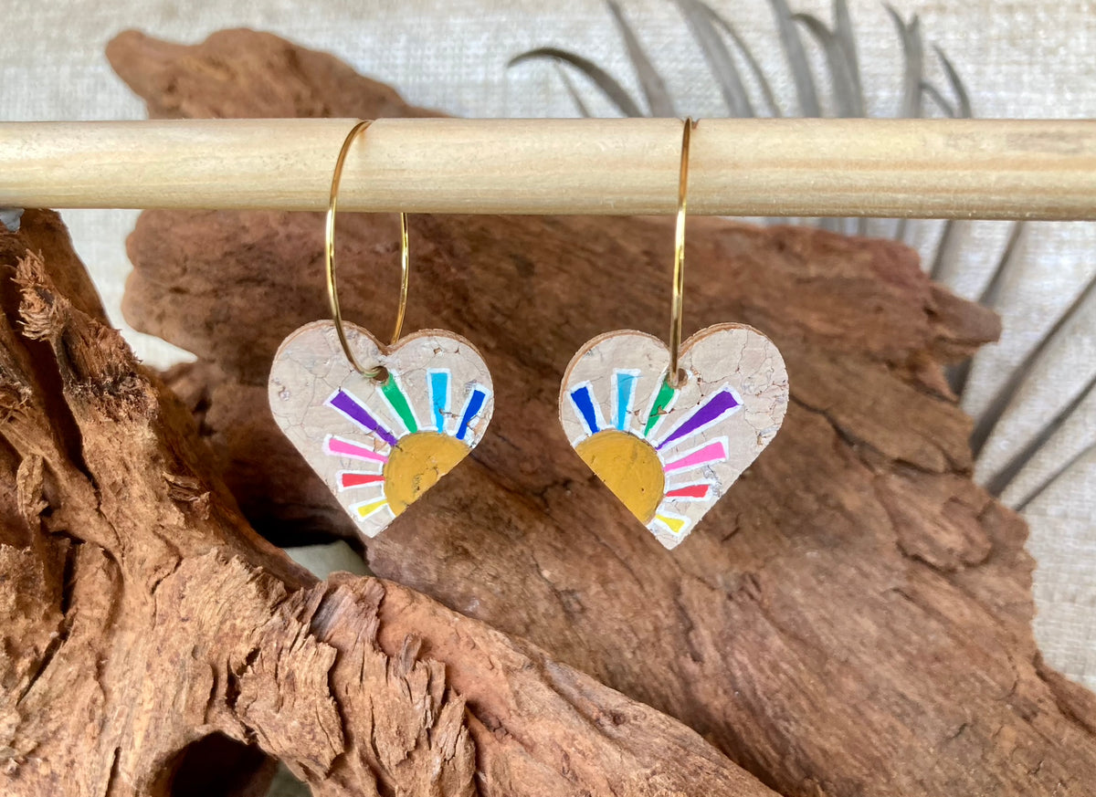 Cork rainbow sun heart earrings on gold hoops, hanging from a wooden dowel with cork bark background
