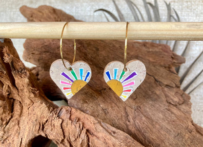 Cork rainbow sun heart earrings on gold hoops, hanging from a wooden dowel with cork bark background