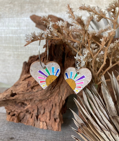 Handpainted rainbow sun heart cork earrings on silver hoops, resting on cork bark with dried botanicals background