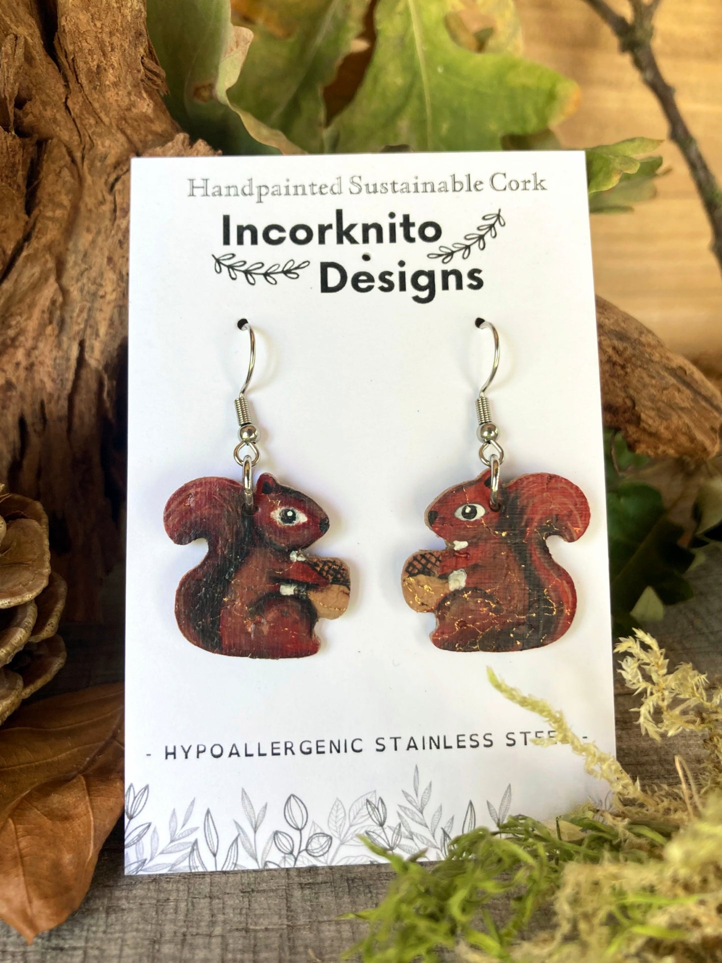 Handpainted red squirrel earrings on Incorknito Designs card surrounded by pine cones and autumn leaves