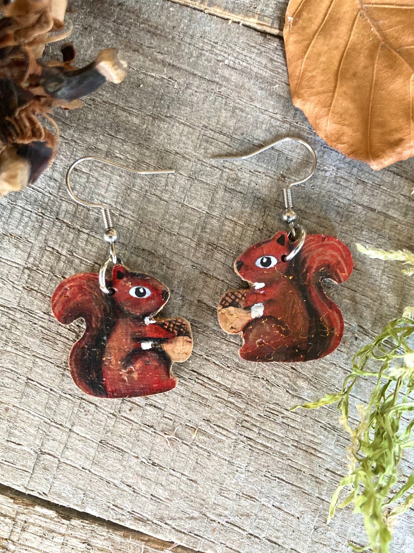 Cork red squirrel earrings flat lay on weathered wood with autumn leaves, pine cone and green moss.