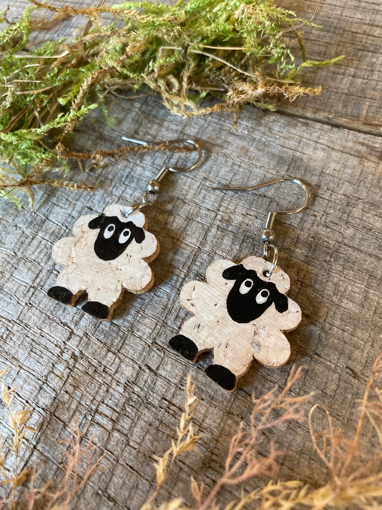 Handpainted cork sheep earrings on silver hooks, flat lay on weathered wood with dried green moss