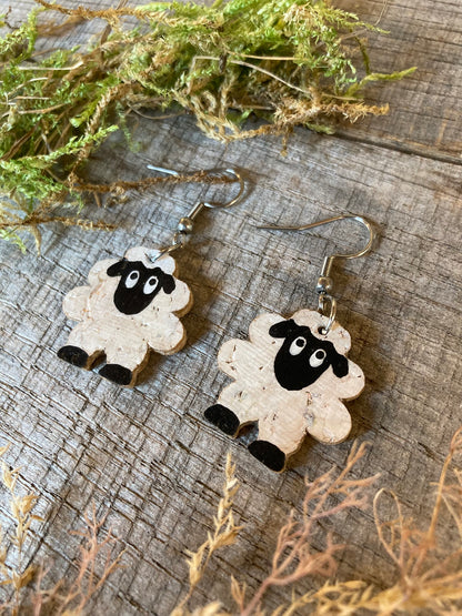 Handpainted cork sheep earrings on silver hooks, flat lay on weathered wood with dried green moss