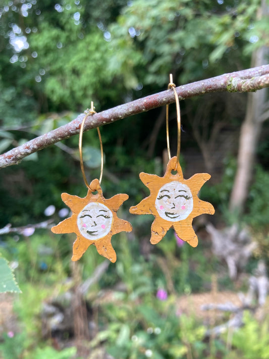 Handpainted cork sun face earrings on gold hoops, hanging from a garden branch with soft green bokeh background