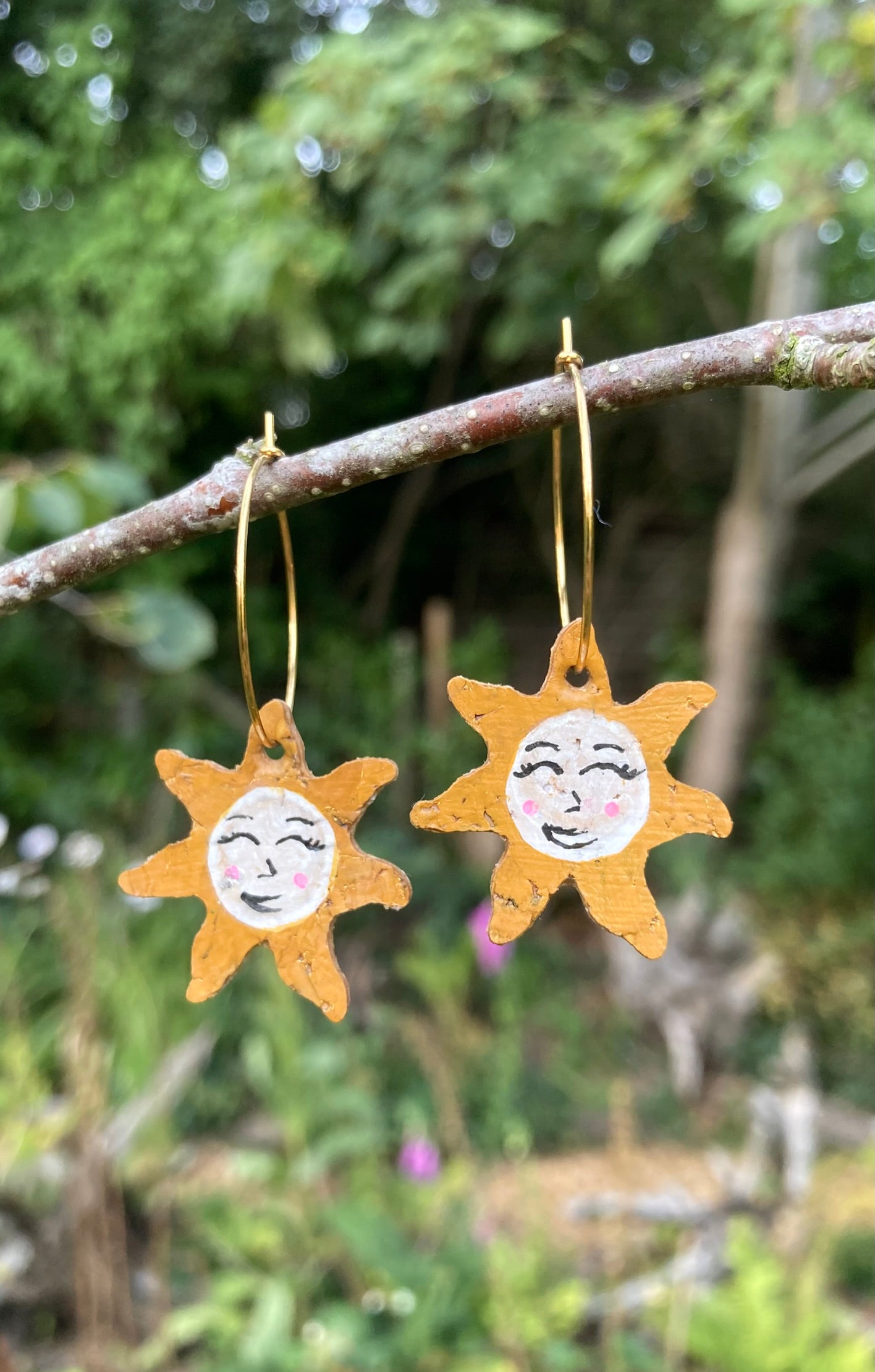 Handpainted cork sun face earrings on gold hoops, hanging from a garden branch showing smiling face detail with natural green background