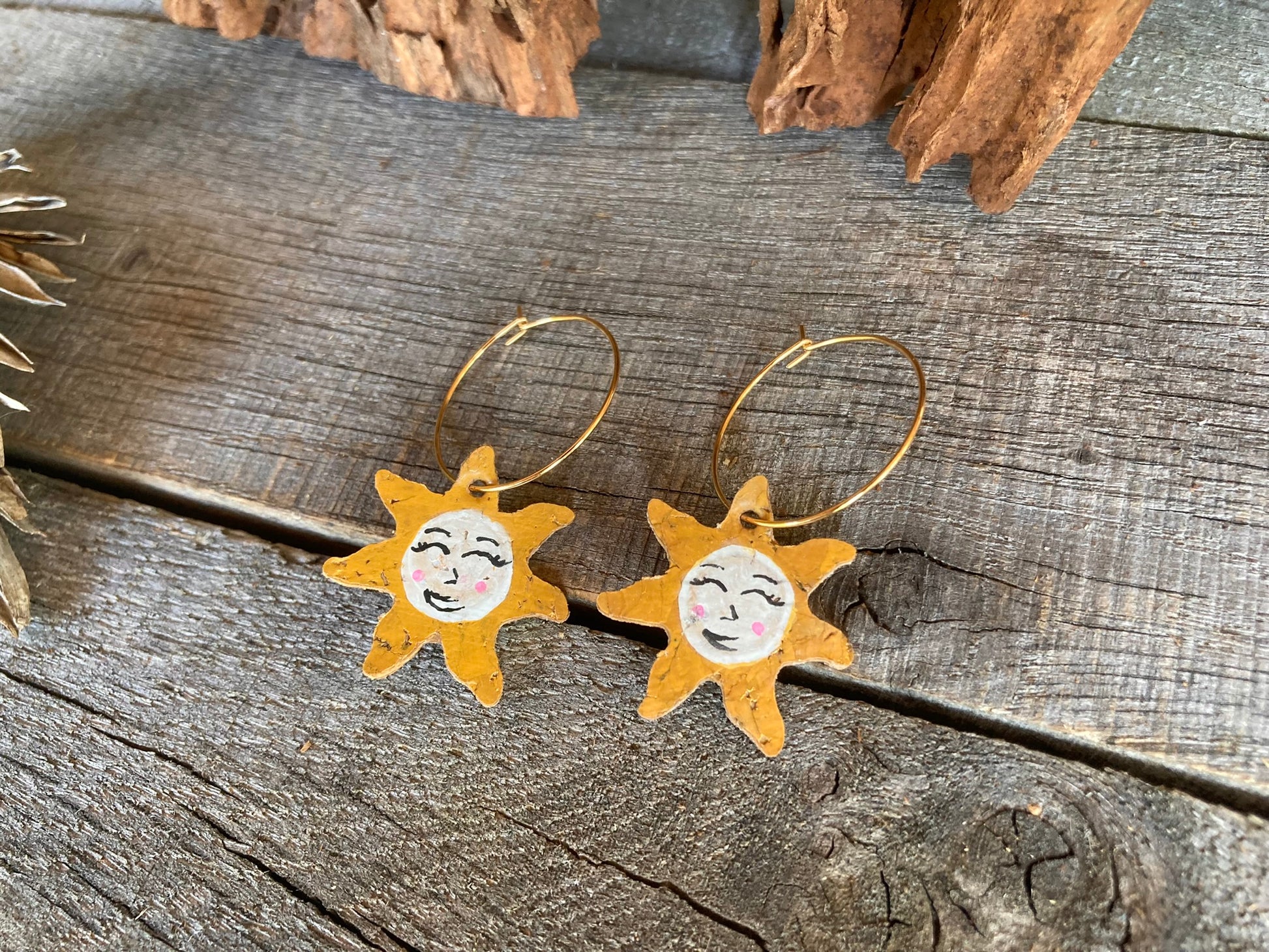Close-up of handpainted cork sun face earrings on gold hoops, flat lay on weathered wood with cork bark background showing smiling face and pink blushed cheeks