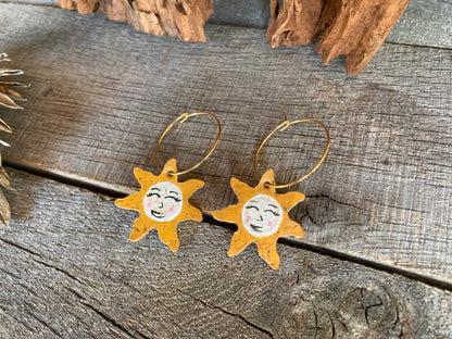 Close-up of handpainted cork sun face earrings on gold hoops, flat lay on weathered wood with cork bark background showing smiling face and pink blushed cheeks
