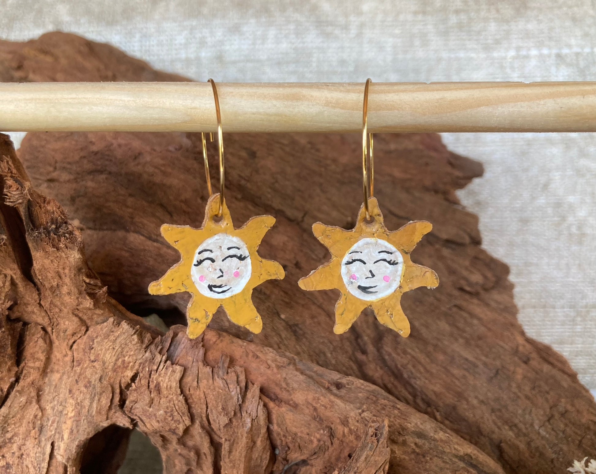 Handpainted cork sun face earrings on gold hoops, hanging from a wooden dowel against cork bark background
