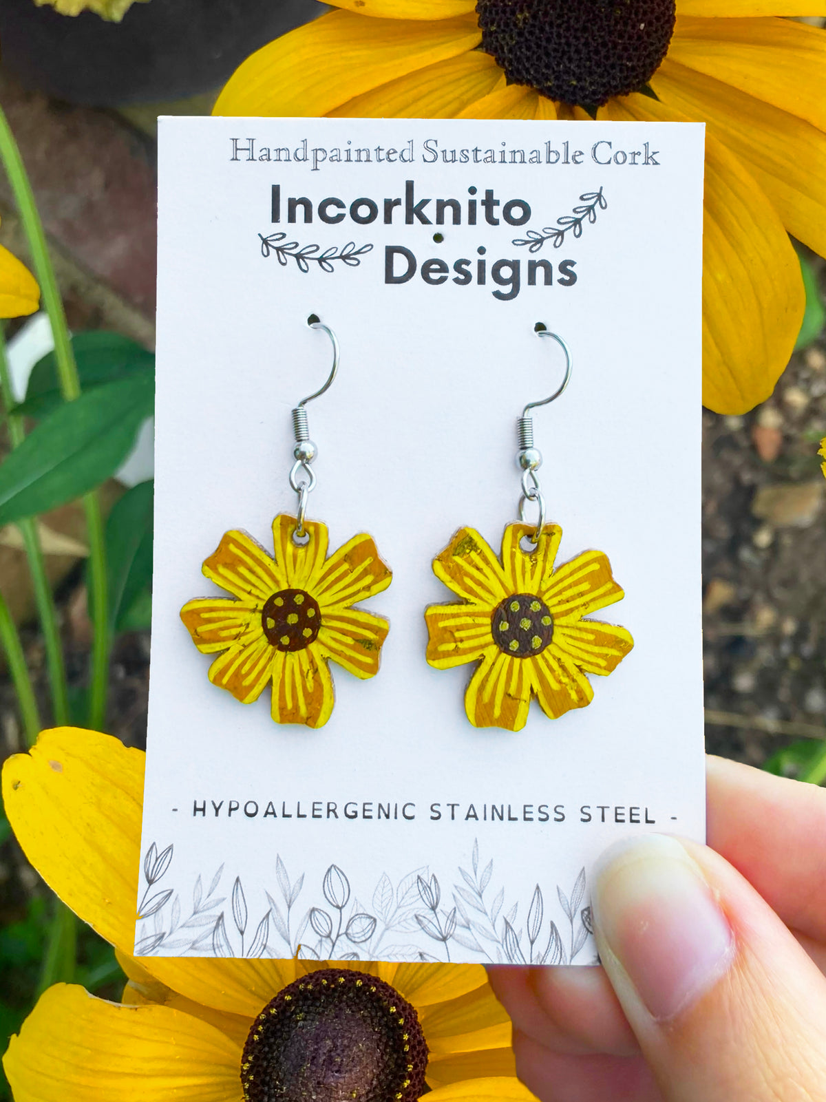 Handpainted cork sunflower earrings on silver hooks, displayed on Incorknito Designs branded card held in hand surrounded by rudbekia