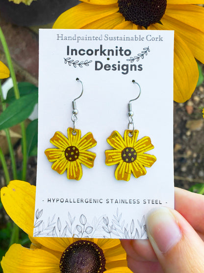 Handpainted cork sunflower earrings on silver hooks, displayed on Incorknito Designs branded card held in hand surrounded by rudbekia