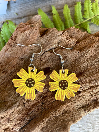Handpainted cork sunflower earrings on silver hooks, resting on cork bark with fern leaves in the background