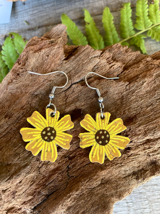 Handpainted cork sunflower earrings on silver hooks, resting on cork bark with fern leaves in the background