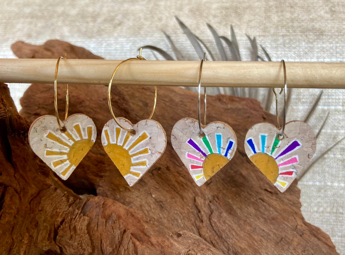 Yellow and rainbow sun heart cork earrings on gold hoops, hanging from a wooden dowel with cork bark background