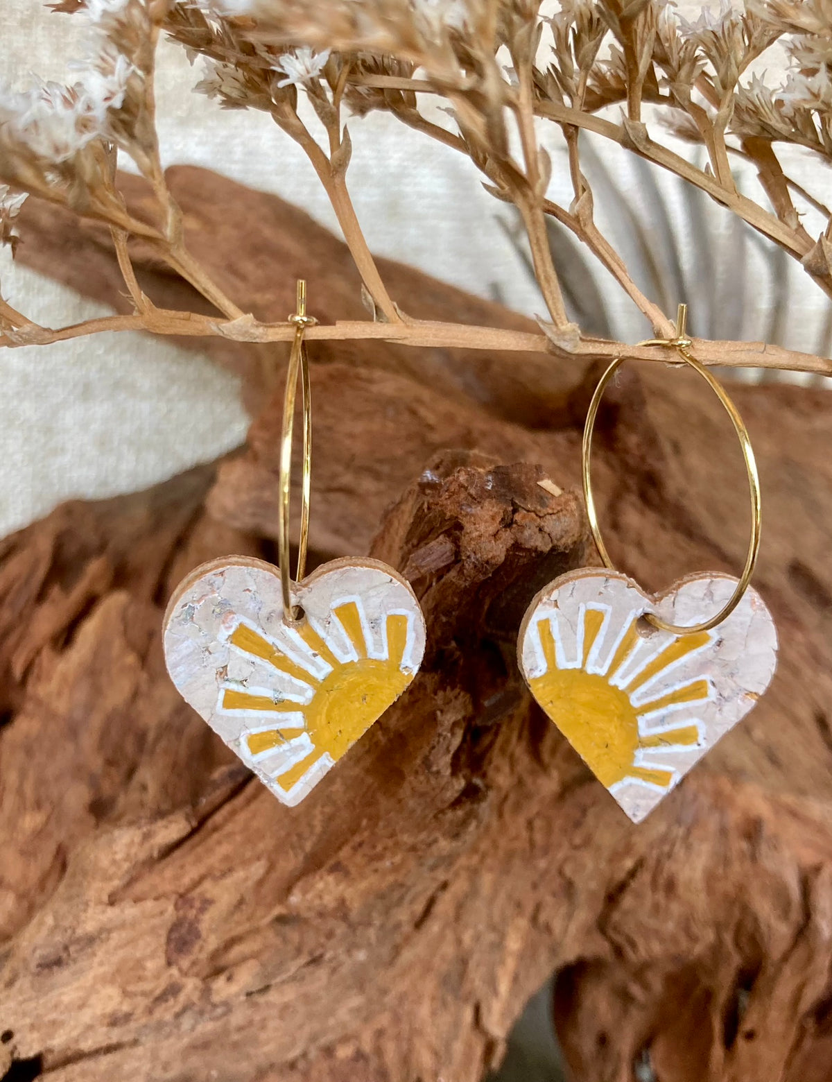 Cork yellow sun heart earrings on gold hoops, hanging from a dried branch with cork bark and dried white botanicals