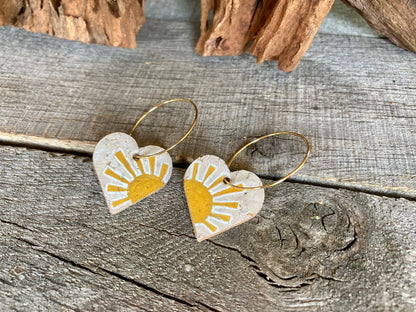 Handpainted yellow sun heart cork earrings on gold hoops, flat lay on weathered wood with cork bark