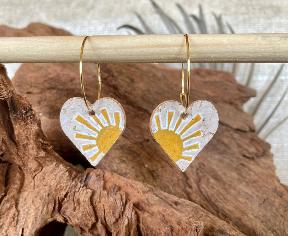 Cork yellow sun heart earrings on gold hoops, hanging from a wooden dowel with cork bark background