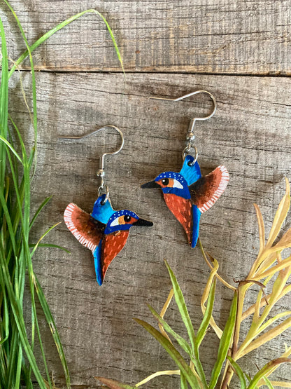 Handpainted blue and orange kingfisher cork earrings on silver hooks flat lay on weathered wood with green and yellow grass