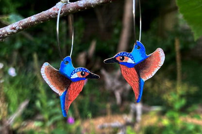 Handpainted kingfisher cork earrings on silver hoops hanging from a branch against green woodland — electric blue and burnt sienna sustainable jewellery