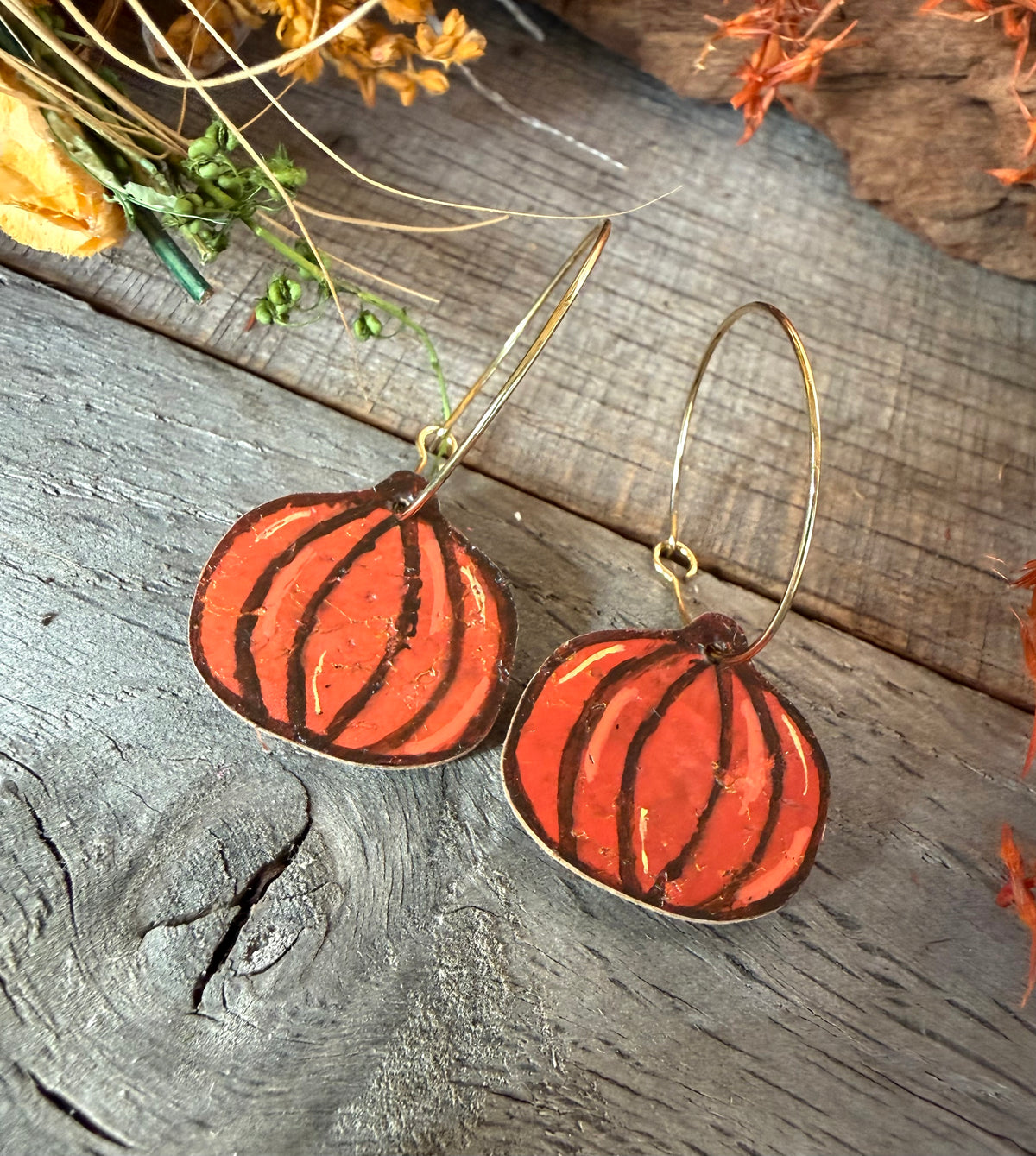 Orange pumpkin cork earrings on gold hoops, showing dark brown segment lines, lighter highlights and gold flecks against weathered wood