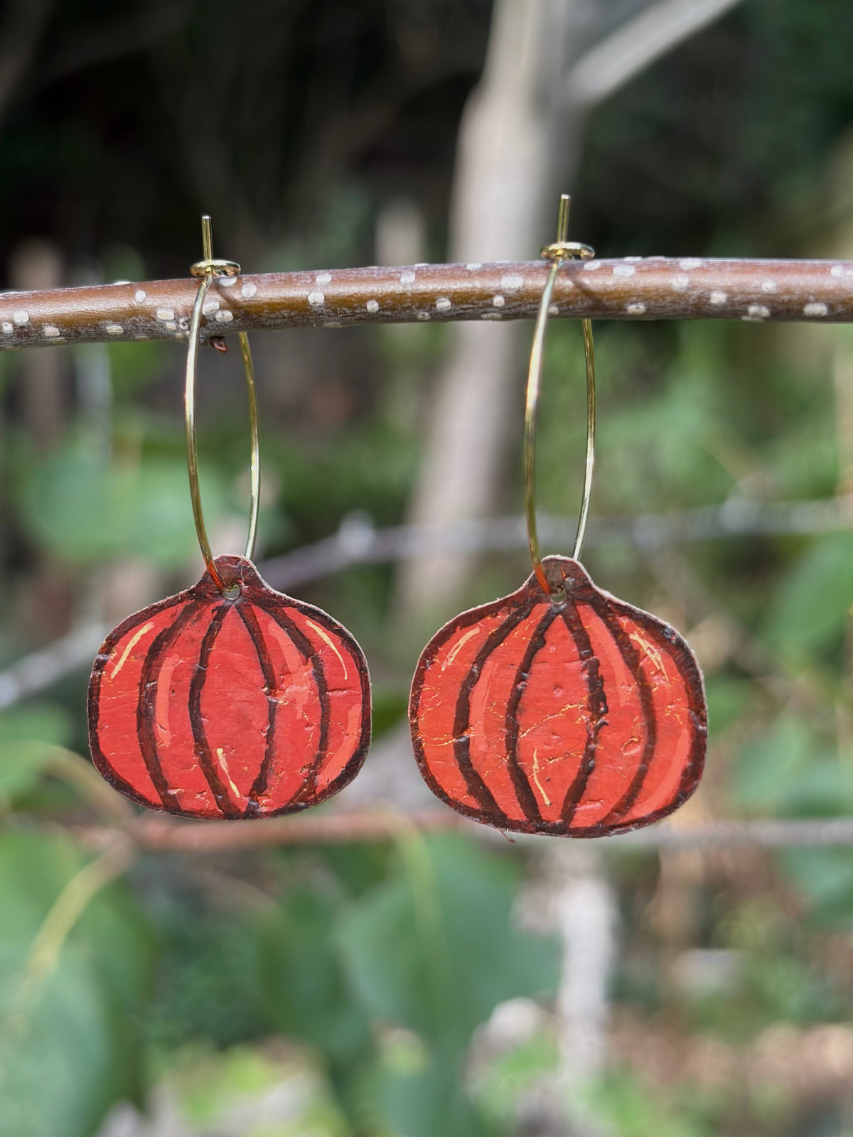 Large handpainted pumpkin earrings suspended from a twig on gold hoops, with a softly blurred green woodland background.