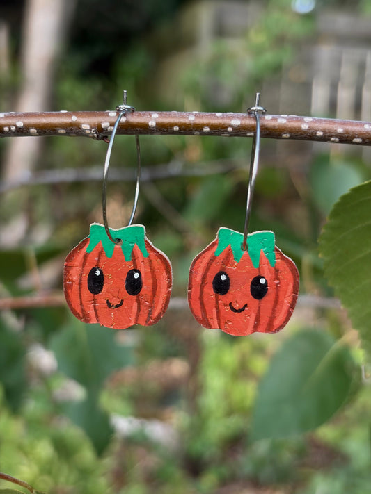 Handpainted large smiley pumpkin cork earrings hanging from a twig on silver hoops, against a softly blurred green background.