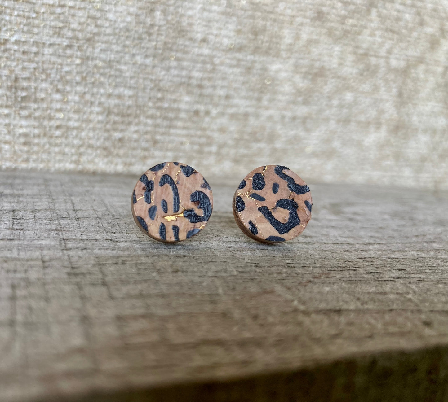 Handpainted leopard print cork stud earrings with black spots and gold fleck with a linen surface background