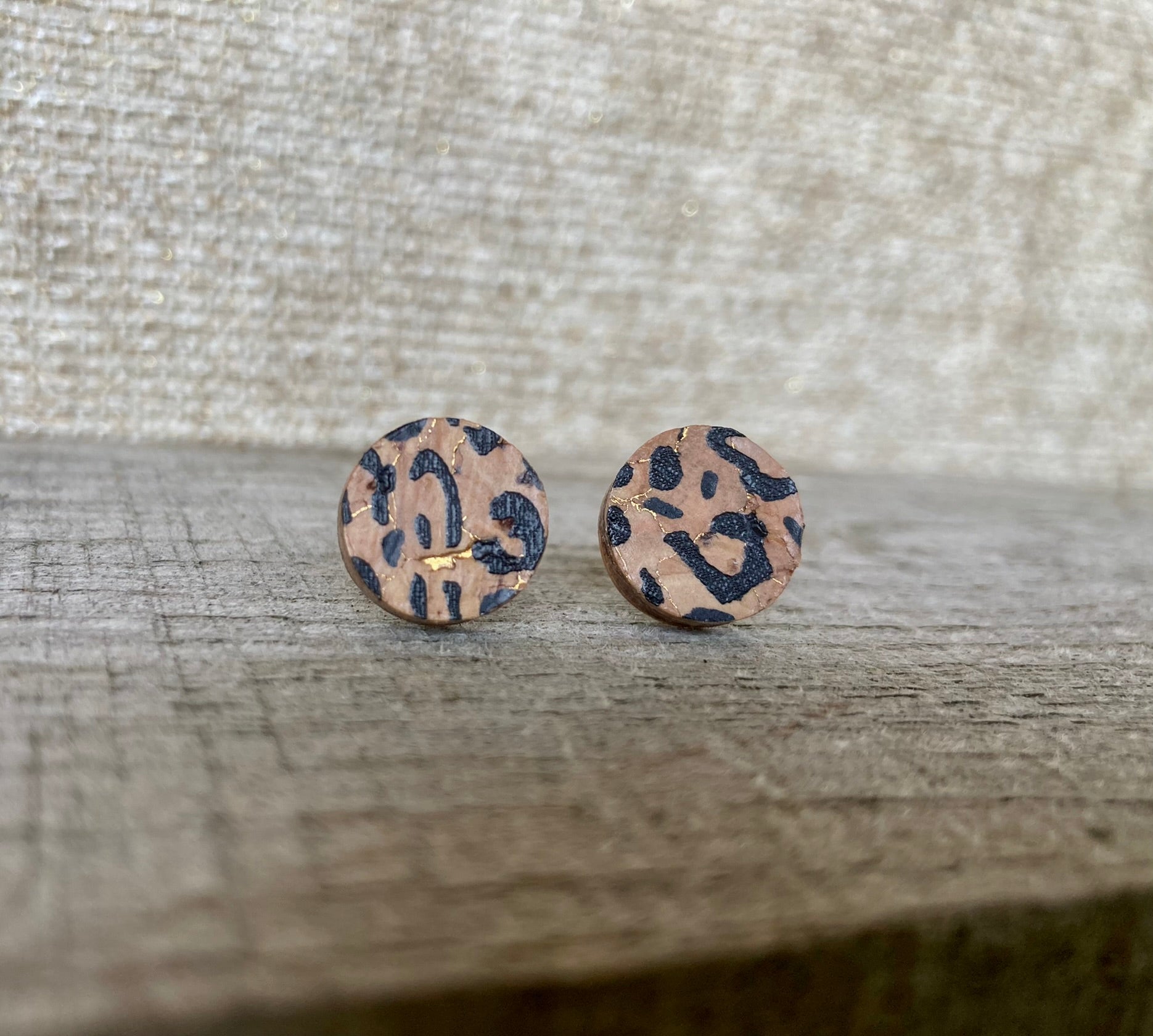 Handpainted leopard print cork stud earrings with black spots and gold fleck with a linen surface background
