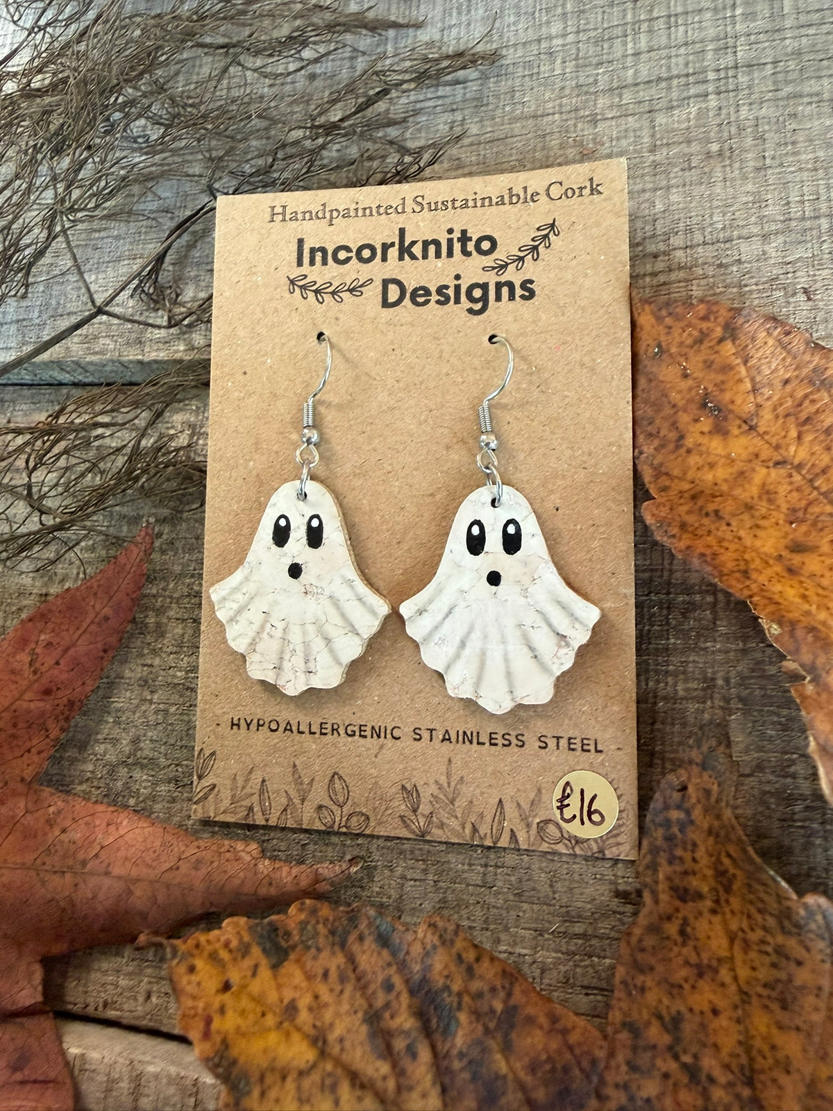 Little Boo ghost earrings with silver-coloured hooks on a kraft display card surrounded by autumn leaves.