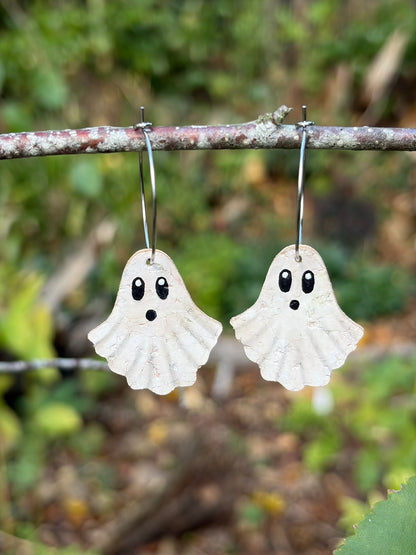 White handpainted cork ghost earrings with surprised faces, hanging from silver-coloured hoops on a woodland twig.