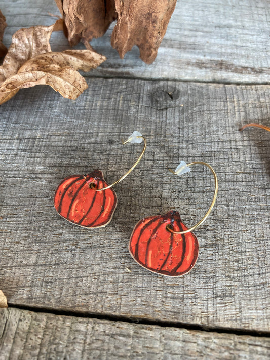 Handpainted orange pumpkin cork earrings on gold open back mini hoops, displayed on weathered wood beside dried leaves.