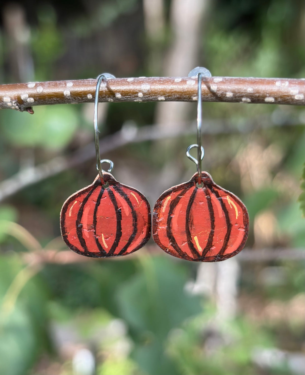 Handmade pumpkin cork earrings suspended from a twig on silver hooks, with orange painted details and gold flecks against a blurred green background.
