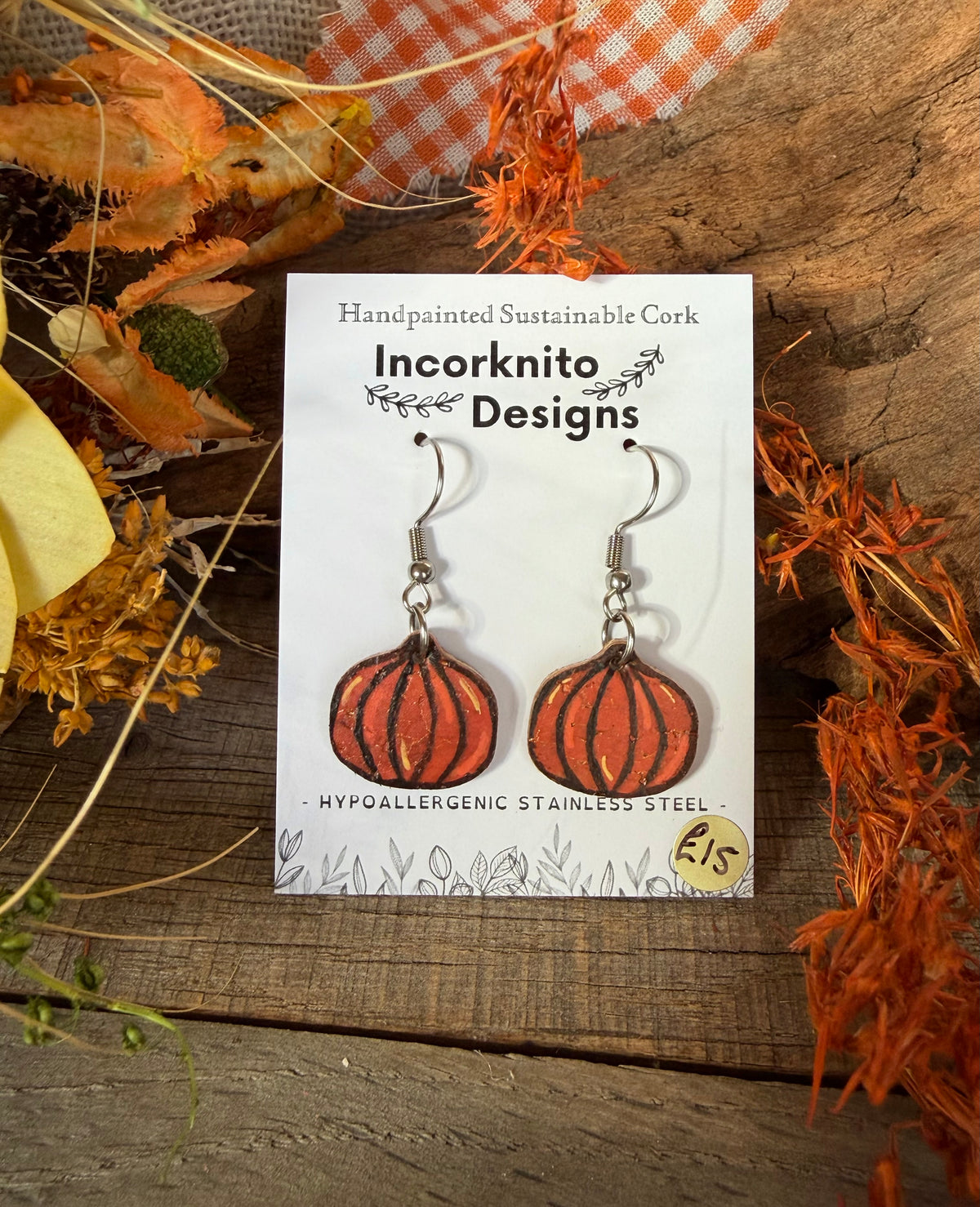 Mini pumpkin cork earrings with silver hooks on an Incorknito Designs card, surrounded by autumn foliage and orange gingham fabric