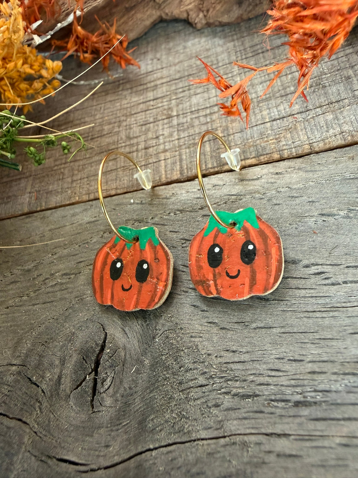 Gold open back hoop earrings with smiling orange pumpkin charms, showing white eye highlights, green tops and the natural gold-flecked cork texture.
