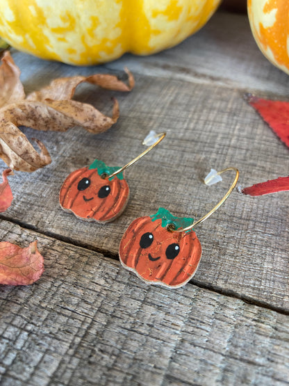 Handpainted smiley pumpkin cork earrings on gold mini hoops, styled on weathered wood with autumn leaves and decorative pumpkins